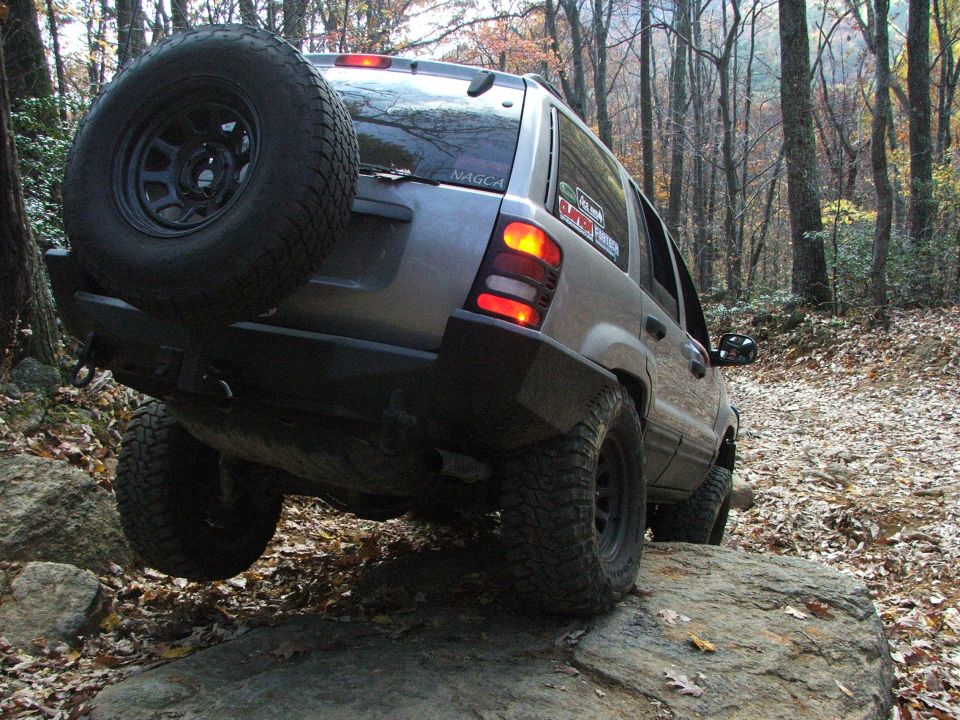 Jeep Grand Cherokee with spare tire on rear bumper climbing over rocks on a wooded trail