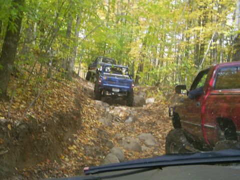 1977 Ford F-150 - Two pickup trucks navigate a challenging, rocky off-road trail through a wooded area with autumn foliage.