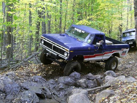 1977 Ford F-150 - A blue 1977 Ford F-150 navigates a rocky trail in a wooded area, with another vehicle visible in the background.