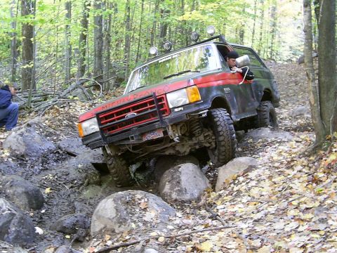 1977 Ford F-150 - Off-road SUV with red grille and black body navigates a challenging rocky path in a wooded area.