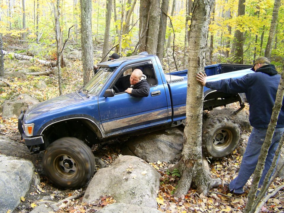 Blue pickup truck navigating a rocky trail in a forest with two people outside