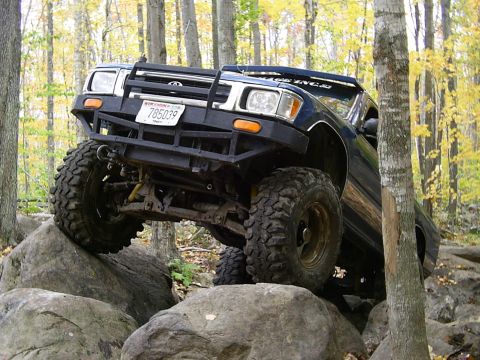 1977 Ford F-150 - A modified Toyota truck tackles a rocky trail in a wooded area.