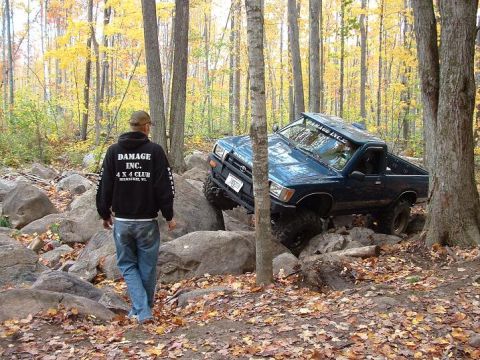1977 Ford F-150 - A blue pickup truck is shown off-roading through a rocky terrain in a wooded area.