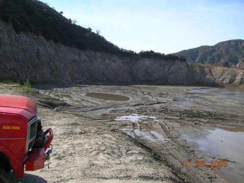 1985 Ford Bronco - The front of a red truck is visible in the foreground of a scene depicting a dry, muddy lake bed with tire tracks and a small pool of water.