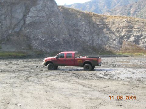 1985 Ford Bronco - A red pickup truck is seen in a muddy, outdoor environment with a rocky hillside in the background.