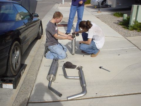 1993 Ford Probe - Two people work on the exhaust system of a black 1993 Ford Probe coupe parked on a driveway.