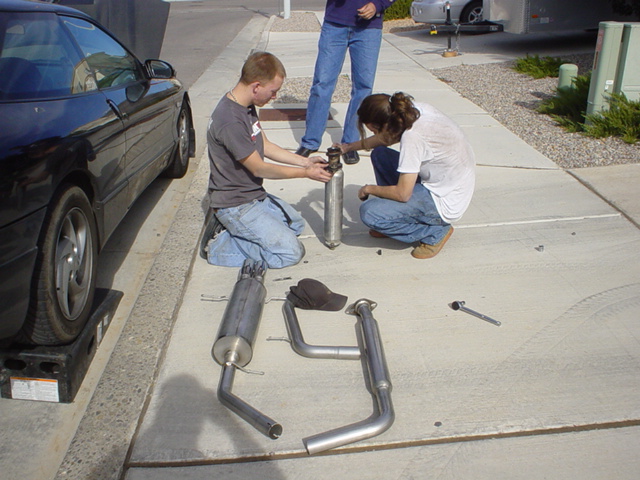 Black 1993 Ford Probe coupe with hood open, parked on a driveway with exhaust system parts laid out on the concrete