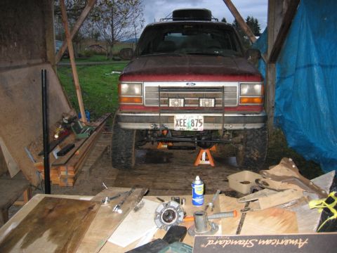 1991 Ford Explorer - Front view of a muddy red Ford Explorer up on orange jack stands under a rough open shed, with the front axle and leaf/radius arm suspensio…