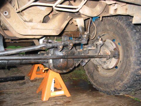 1991 Ford Explorer - Underbody shot of a solid front axle supported on two orange jack stands over a plank floor, showing the differential housing, tie rod and…