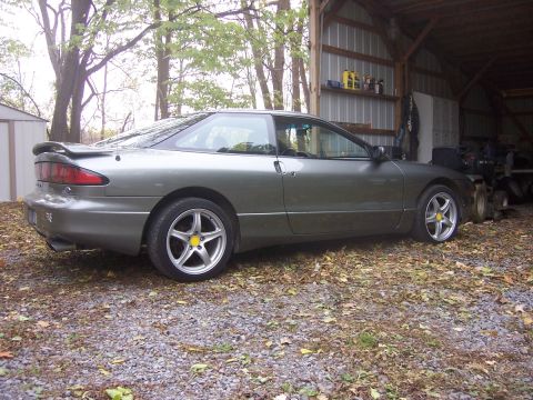 1996 Ford Probe - A gray coupe sits on a gravel surface next to a shed and a barn with equipment inside.