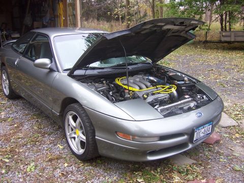 1996 Ford Probe - Silver two-door hatchback coupe parked on gravel outside a garage with its hood propped open, showing a transverse V6 with bright yellow af…