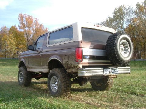 1993 Ford Bronco - A muddy, lifted Ford Bronco is parked on a grassy field with trees in the background.