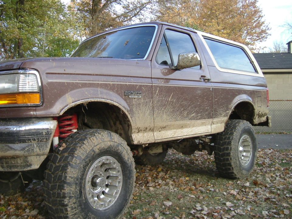 Muddy lifted tan and brown Ford Bronco with red coil springs and big mud tires, left front view