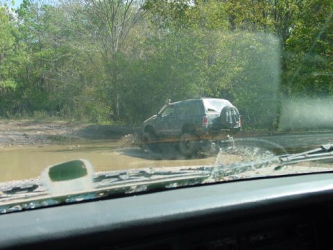 1992 Chevrolet S10 - A dark SUV navigates a muddy off-road path, kicking up water as it goes.