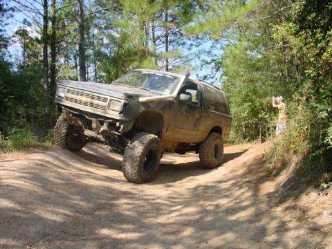 1992 Chevrolet S10 - A lifted, mud-covered Chevrolet Blazer K5 navigates a wooded trail with a person in the background.