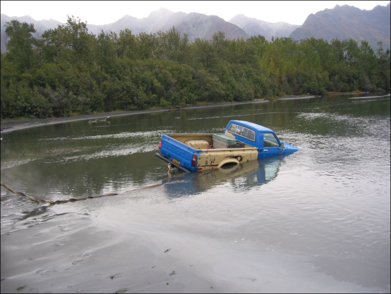 Blue Toyota-style mini pickup half-submerged in a shallow river with mountains behind