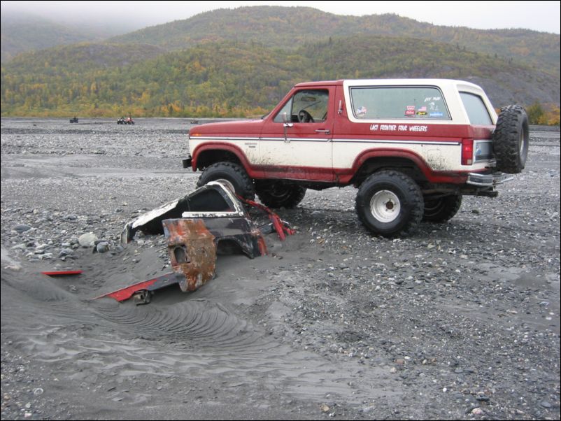 Lifted red and white full-size Bronco parked on gray river gravel beside a wrecked body shell