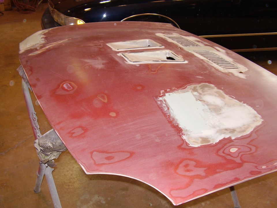 Car hood being prepped for paint, with body filler patches and vent cutouts. Another car is visible in the background