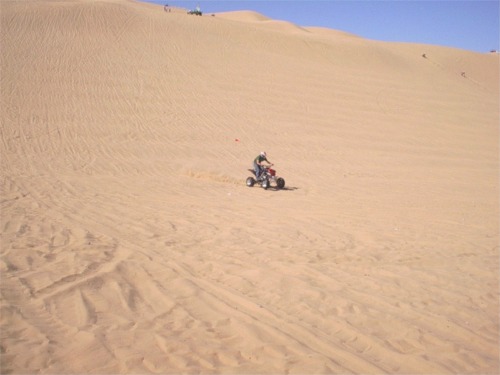 ATV rider on a sand dune, with other people and vehicles visible further up the dune