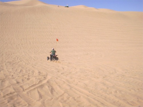 ATV with red flag rides down a sand dune under a blue sky
