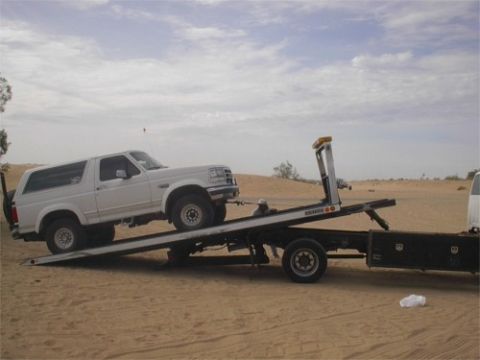 1993 Ford Bronco - A white 1993 Ford Bronco is loaded onto a tow truck in a sandy, desert environment under a cloudy sky.