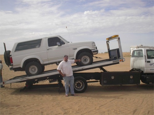 White 1993 Ford Bronco loaded onto a tow truck with a man standing next to it