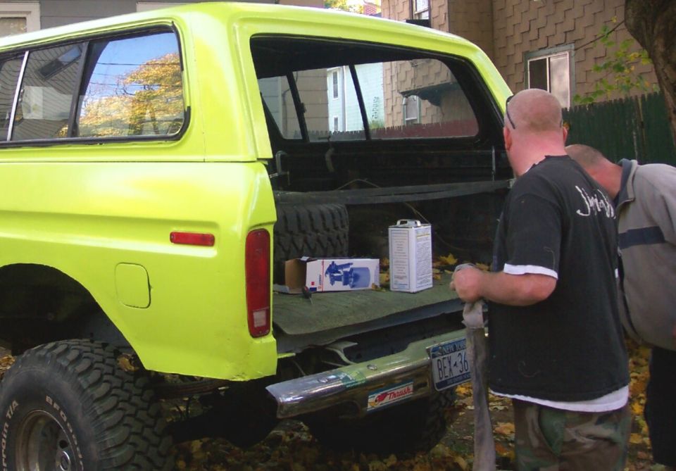 Yellow 1978 Ford Bronco rear view with tailgate open, showing spare tire and supplies. Two men are looking into the bed