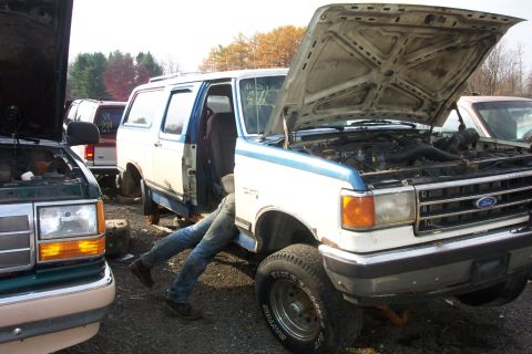 1978 Ford Bronco - A person works on a white and blue Ford Bronco in a junkyard setting with other vehicles visible.