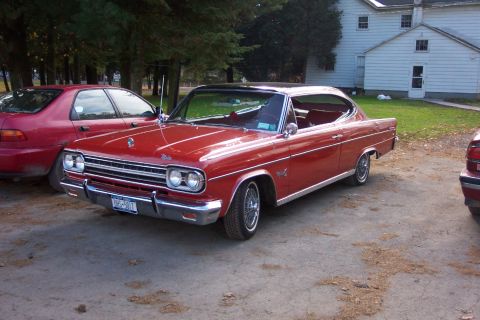 1978 Ford Bronco - A red 1965 AMC Rambler American coupe is parked outdoors next to another red vehicle.