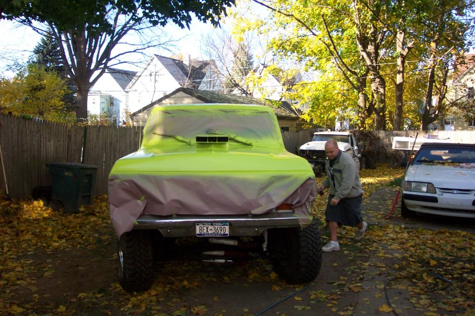 Rear view of a Bronco covered in green plastic, with a man nearby and another car parked to the right. License plate reads BEX-3690