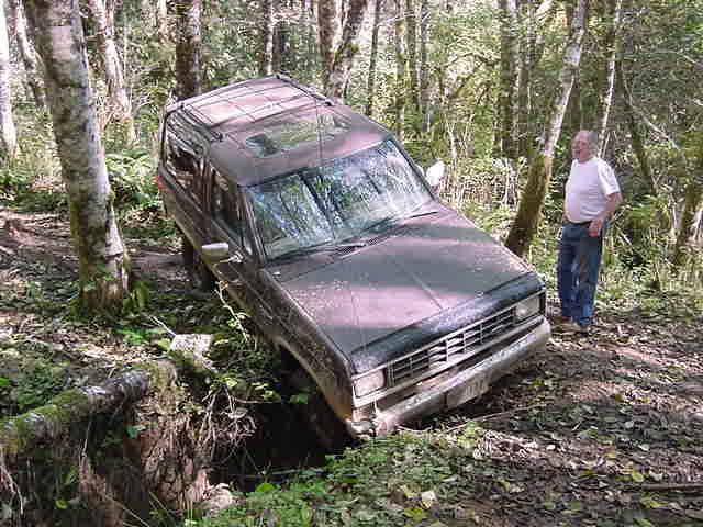 Muddy dark green Ford Bronco II nose-down in a ditch on a forest trail, man standing beside it