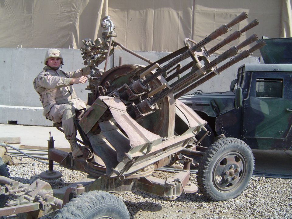 Soldier sits on a mounted Gatling gun with a military truck in the background