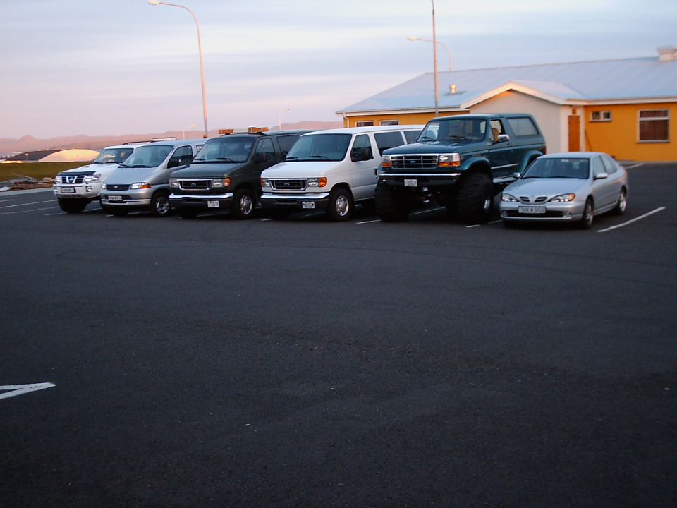 Lifted teal Ford Bronco on large tires parked in a row of vehicles at dusk.