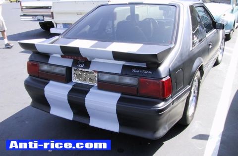 1986 Ford Bronco - A 3/4-rear view of a flat-black Fox-body Mustang hatchback wearing white racing stripes over the bumper and wing, with a Honda emblem and "…