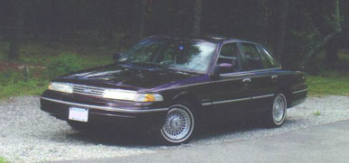 1994 Ford Crown Victoria - A dark-colored sedan is parked on a gravel surface with trees in the background.