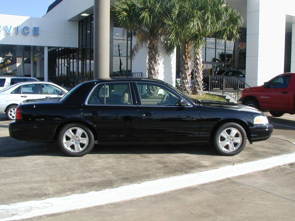 Black 2000 Ford Crown Victoria parked outside a building with palm trees. Another car is visible to the left, and a red truck to the right