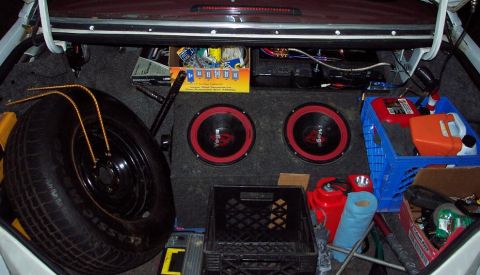 1985 Ford LTD Crown Victoria - The trunk of a car contains a spare tire, audio equipment, and various supplies organized in crates.