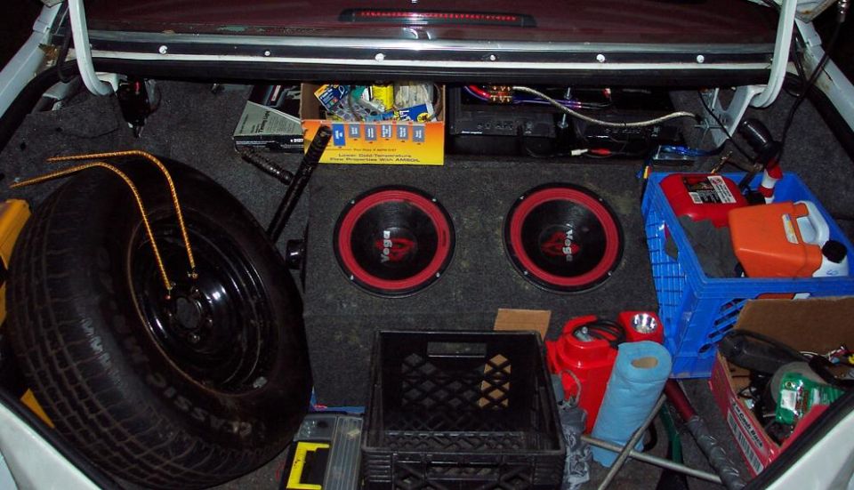 Car trunk interior with spare tire, two subwoofers, tool, and storage crates filled with items