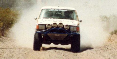 1990 Ford Bronco - white off-road race truck with four roof-mounted driving lights and a blue tube bumper catches air on a dusty desert trail.