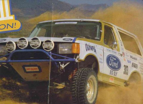 1990 Ford Bronco - white Ford Bronco off-road race truck numbered 303 with Hella lights and Ford Big Oly livery, kicking up dust in the desert.