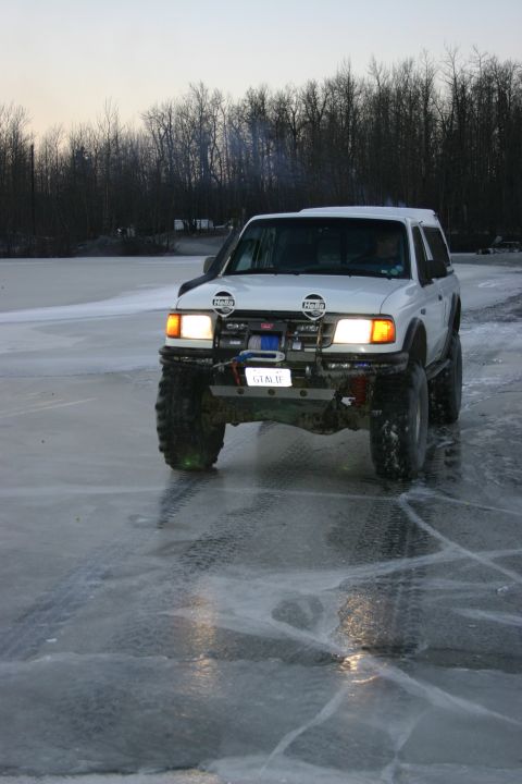 2003 Jeep TJ - White SUV with aftermarket modifications drives on a frozen surface, showing its front end and large tires.