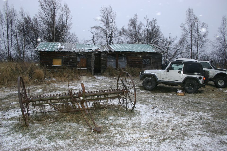 White 2003 Jeep TJ and another SUV parked near a rundown cabin with an old farm rake in the foreground