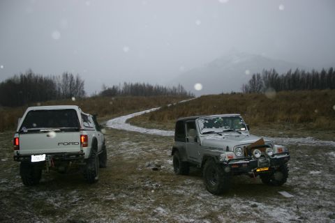 2003 Jeep TJ - Two off-road vehicles, a Jeep Wrangler and a Ford pickup, are parked on a snowy dirt road with a mountain in the background.