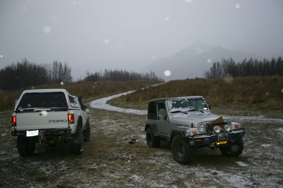 Silver Jeep Wrangler with a black top and a white Ford pickup truck with a camper shell parked on a snowy dirt road