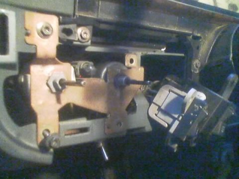 1990 Ford Bronco - Close-up of a disassembled headlight or dash switch mechanism with copper brackets and a control rod behind a vehicle dashboard.