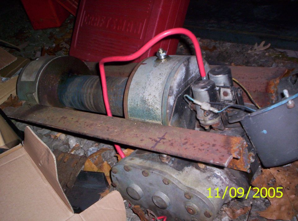 Winch mechanism with red hoses and electrical components, resting on rusty metal