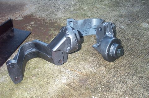 1995 Ford Bronco - Two freshly painted or coated cast steel suspension/axle brackets sitting on a concrete floor, one with a large hex bolt head visible and a…