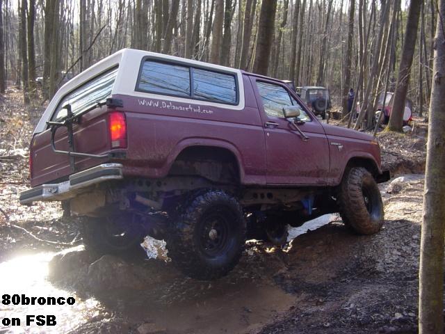 Maroon lifted Ford Bronco with white top crawling over rocks on a muddy forest trail