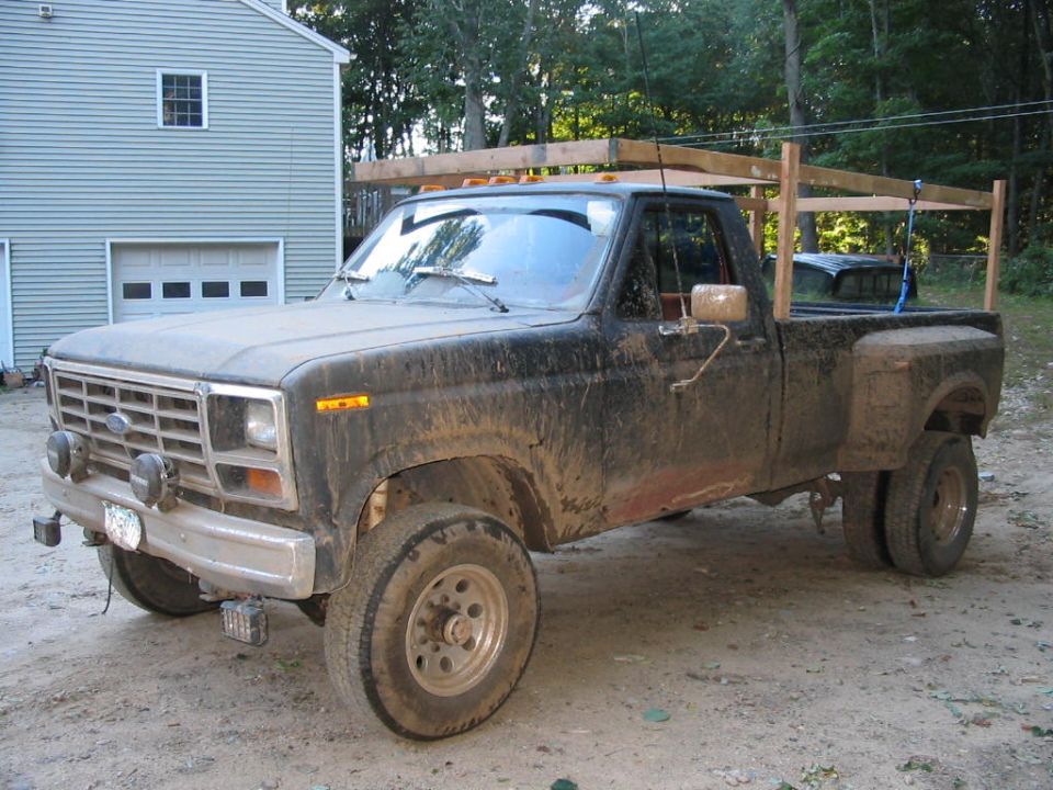 Mud-covered 1980s Ford dually pickup with wooden bed rack and round driving lights on dirt drive