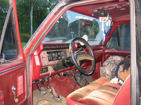 1985 Ford F-350 - The interior of a red pickup truck shows a cracked windshield and worn seats, with camouflage bags on the passenger side.